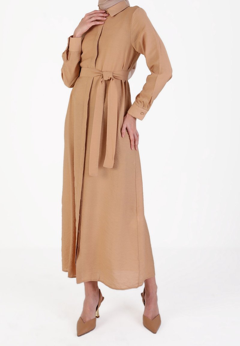 Kvinna dragen i en beige långärmad klänning med bälte, matchande högklackade skor och en beige huvudduk, som står mot en vit bakgrund.