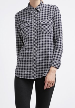 Femme portant une chemise à carreaux noire et grise à boutons avec deux poches poitrine, associée à un pantalon noir, debout devant un fond blanc.