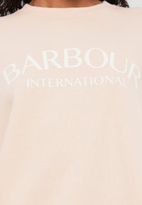 Roosa pusak, millel on valge tekstiga "BARBOUR INTERNATIONAL". Ümmargune kaelus, pehme tekstuur ja sile kangas, mis on mugava lõikega.