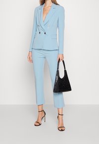 Jasnoniebieski dwurzędowy garnitur z dopasowaną marynarką i skróconymi spodniami, zestawiony z czarną torebką z tłoczeniem imitującym skórę krokodyla oraz czarnymi sandałkami na paseczki.