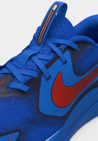 Lähivõte sinisest Nike Cosmic Runneri tossust, millel on punaste triipudega Swoosh ja sinised paelad võrkmaterjalist peal.