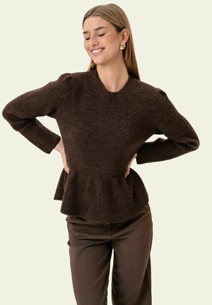 Kvinde smilende med hænderne på hofterne iført en mørkebrun ribbet strikket peplumtrøje og brune bukser på en lys baggrund.