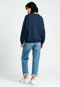 Mornarsko plavi dukserica s opuštenim krojem, rebrastim.manžetama i okruglim izrezom, u kombinaciji s svijetloplavim jeansom s podignutim nogavicama i bijelim tenisicama.