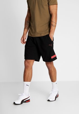 Schwarze Shorts mit rotem Akzent und Puma-Logo, kombiniert mit einem grünen T-Shirt, weißen und schwarzen Turnschuhen sowie weißen Socken.