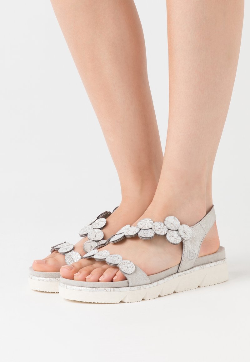 Pieds portant des sandales argent-grises avec des ornements floraux circulaires et des semelles blanches texturées sur un fond blanc.