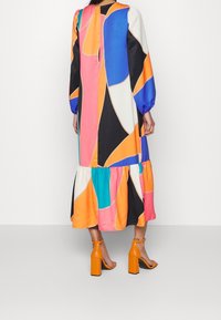 Femme portant une longue robe multicolore à motif abstrait avec des manches bouffantes et des sandales à talons hauts orange, debout contre un mur blanc.