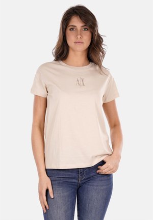 T-shirt in cotone beige con maniche corte, caratterizzata da un logo metallico "AX" centrato sul petto, abbinata a jeans in denim blu.