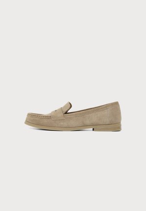 Mocassin plat en daim beige avec petit talon empilé et détail d’une bride penny, montré de profil sur un fond clair uni.