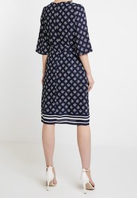 Marinblå klänning med vita geometriska mönster och trekvartsärmar. Har en randig nederkant. Materialet verkar lätt och har en avslappnad passform.