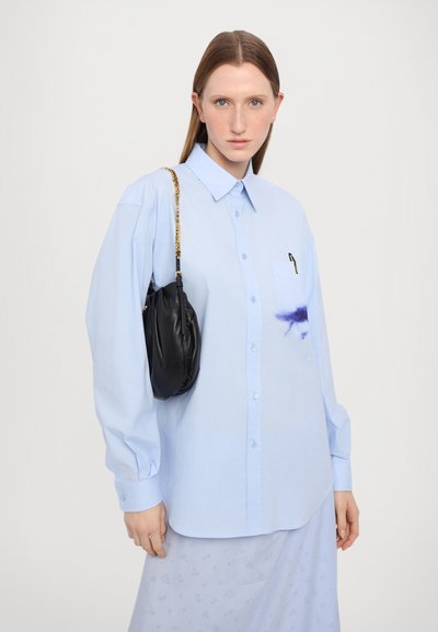 Camicia azzurro chiaro con maniche lunghe e tasca, caratterizzata da un design astratto viola; abbinata a una piccola borsa nera in pelle.