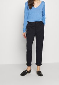 Blauer V-Ausschnitt-Pullover, leichtes Material, lässige Passform. Dunkle Hose, gerade Beinform, am Knöchel umgeschlagen. Schwarze Slipper mit silbernen Akzenten.