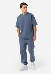 Šviesiai mėlyna medvilninė trumpa rankovė t-shirt su atitinkamomis sportinėmis kelnėmis. Abu drabužiai turi laisvą siluetą ir subtilią logotipo detalę. Baltos sportbačiai papildo aprangą.