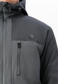 Záběr zblízka na osobu v tmavě šedé bundě s kapucí, která má na hrudi zipovou kapsu a na levé straně nášivku s logem Rip Curl.