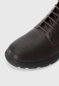 Botín de cuero marrón con puntera redondeada, que presenta cordones oscuros y una superficie texturizada. La suela es de goma negra con un patrón estriado para mayor tracción.