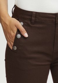 Un pantalon en denim marron présente une coupe ajustée, une poche latérale avec des accents en métal, et une texture lisse, associé à un haut blanc.