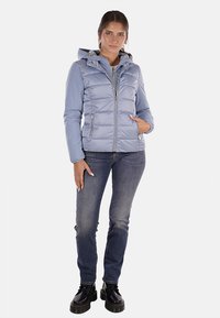 Giacca imbottita azzurro chiaro con cappuccio, zip argentata e due tasche frontali, indossata sopra jeans scuri e stivali neri. Texture liscia, design casual.