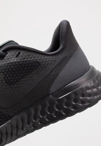 Zapatilla deportiva negra con parte superior de malla, acentos lisos y suela acolchada con textura ondulada. Logo visible en el lateral.