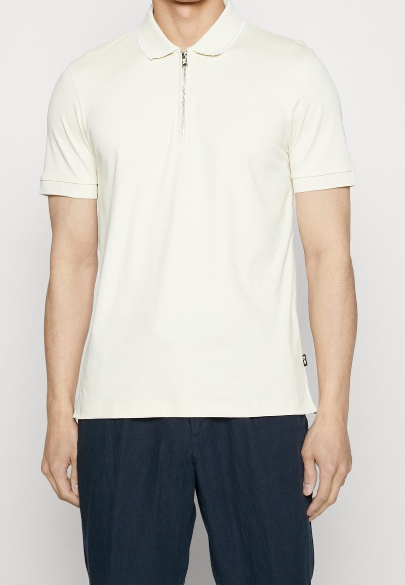 Homme portant un polo blanc uni à manches courtes avec une fermeture éclair frontale, assorti à un pantalon bleu marine foncé, debout sur un fond blanc.