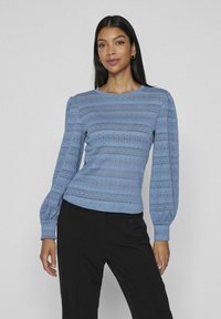 Světle modrá blůza s dlouhými rukávy, texturovanými vertikálními pruhy a nabíranými rukávy. Má kulatý výstřih a přiléhavý lem.