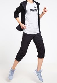 Czarna sportowa kurtka na szarej koszulce Puma, zestawiona z czarnymi spodenkami capri i jasnoniebieskimi trampkami. Kurtka zapinana na zamek z przodu.