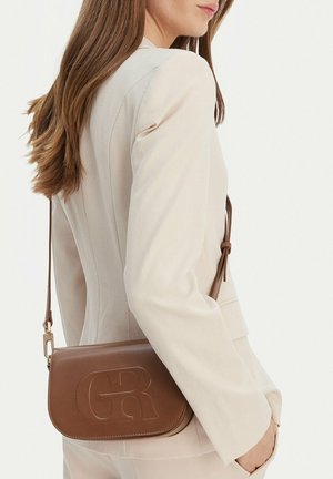 Kobieta w beżowej marynarce niosąca brązową skórzaną torbę na ramię z wytłoczonym logo "GR" po bokach.