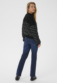Woman with long blonde hair wearing black striped sweater, blue jeans, and black shoes, standing and looking over her shoulder against a white background.