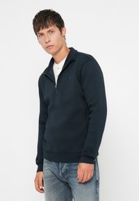 Pull-over sweat-shirt bleu marine à demi-zip avec une texture douce, doté d'un col montant et d'un petit logo sur la poitrine. Associé à un jean clair.