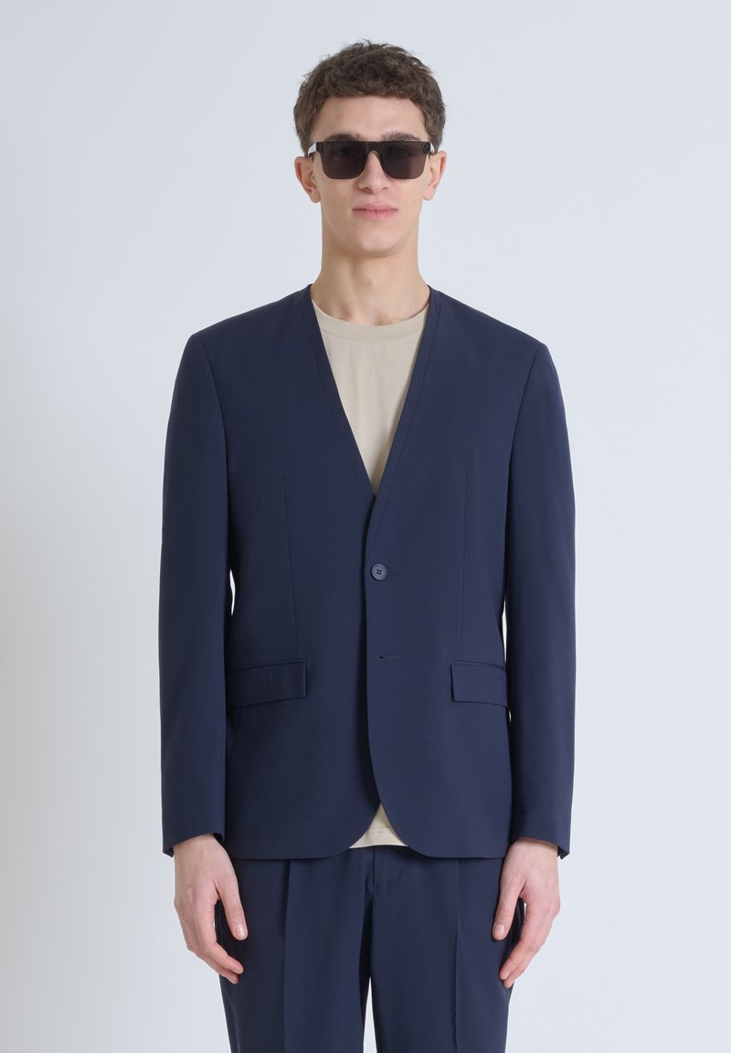 Blazer blu navy con colletto a punta, chiusura con un solo bottone e due tasche frontali, abbinato a una t-shirt beige a girocollo sotto.