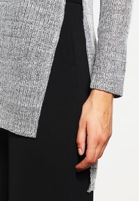Maglia grigia a maglia testurizzata con maniche lunghe, caratterizzata da uno spacco laterale, abbinata a pantaloni neri, che mette in risalto una silhouette casual e un contrasto di tessuti.