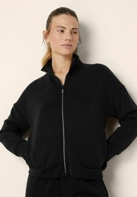 Felpa nera con zip e colletto alto, caratterizzata da una texture liscia e una zip decorativa. Design loose-fit con maniche lunghe.