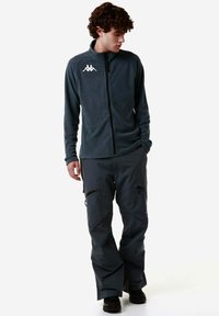 Giacca in pile grigia scura con colletto alto e logo bianco, abbinata a pantaloni impermeabili grigi scuri con tasche con zip.