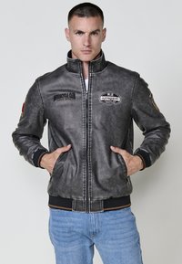Chaqueta de motocicleta de cuero gris con un cuello alto, bolsillos frontales y varios parches bordados, que presenta una cintura y puños acanalados.