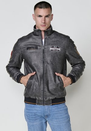 Chaqueta de motocicleta de cuero gris con un cuello alto, bolsillos frontales y varios parches bordados, que presenta una cintura y puños acanalados.