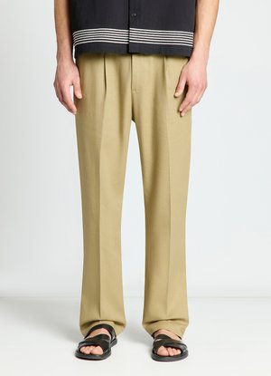 Homme portant un pantalon beige plissé, des sandales noires à bouts ouverts et une chemise noire avec des rayures blanches à l'ourlet, debout devant un fond uni.