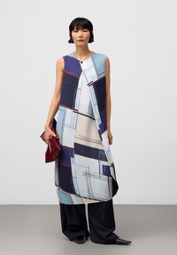 Vrouw in een mouwloze tuniek met geometrisch patroon, die over zwarte wijde broek draagt, met een opgevouwen bordeauxrode clutch in de hand, staand tegen een effen achtergrond.
