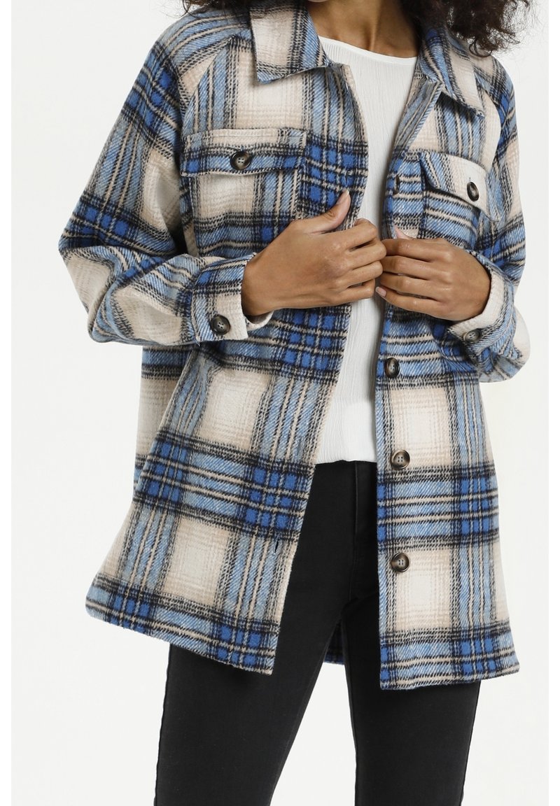 Personne portant une veste à carreaux bleue, beige et noire boutonnée sur un haut blanc et un pantalon noir, ajustant la veste près de la poitrine.