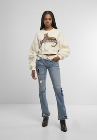 Sweatshirt court crème avec un motif léopard, associé à un jean déchiré bleu clair avec des accents de déchirure et un ourlet effiloché, porté avec des bottes noires.