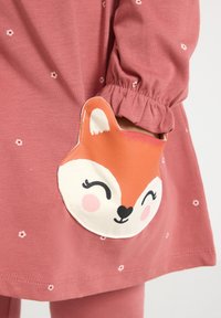 Das rosa Kleid eines Kindes mit kleinen Blumenmustern und einer Tasche in Form eines Fuchsgesichts, dessen Hand teilweise in die Tasche gesteckt ist.