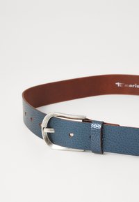 Cintura in pelle blu con texture, dotata di fibbia argentata e numerosi fori, poggiata su una superficie bianca.
