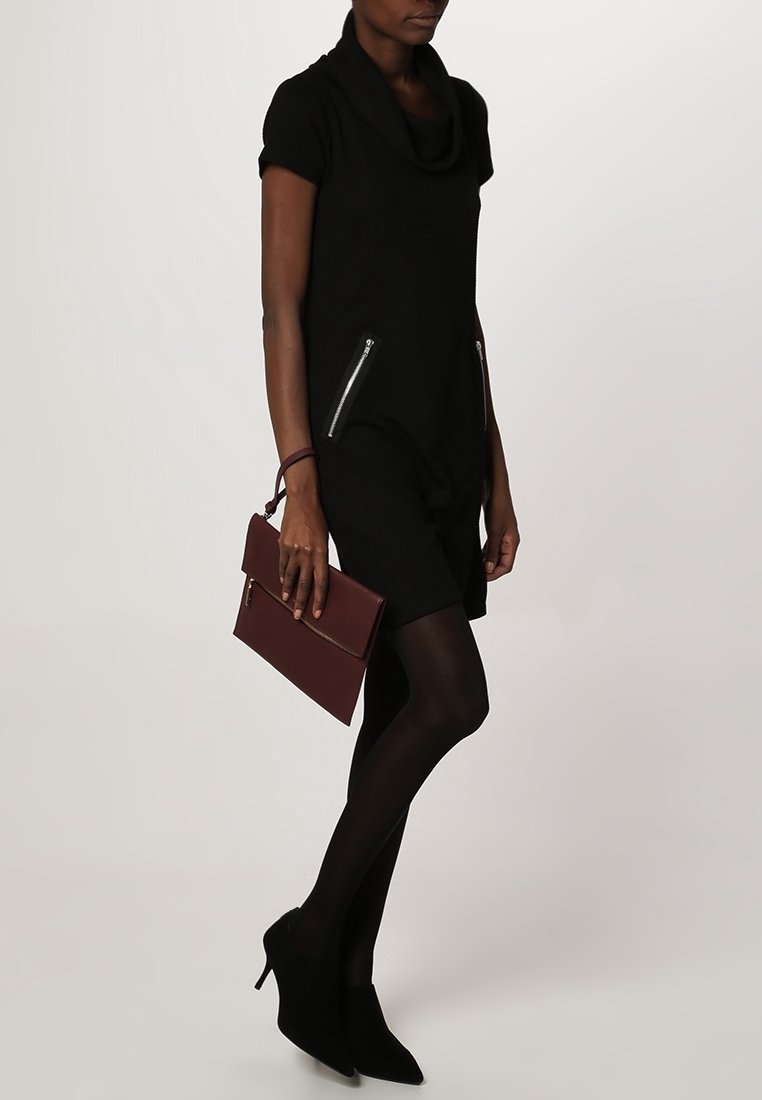 Robe noire à col cheminée avec manches courtes, dotée de deux poches zippées. Le modèle tient une pochette bordeaux, associée à des escarpins noirs et des collants.