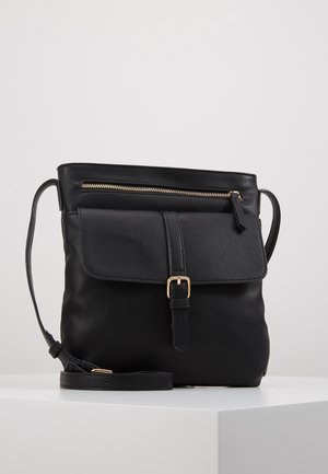 Sac bandoulière en cuir noir avec rabat avant fermé par une boucle dorée et une poche zippée sur le dessus, présenté sur une surface blanche.