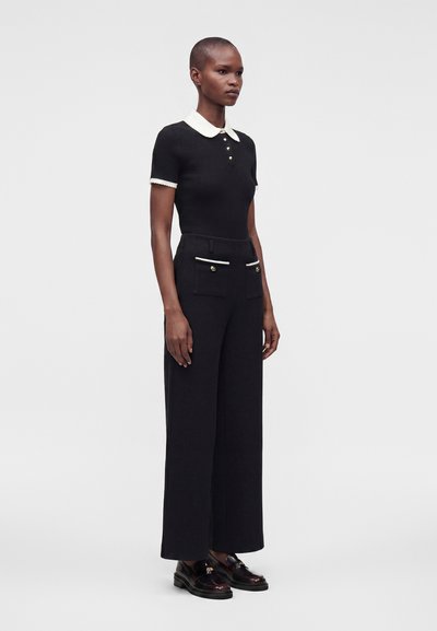 Femme portant un haut noir à manches courtes avec col blanc, un pantalon noir à jambes larges avec poches avant et des mocassins noirs, debout de profil.