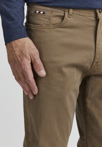 Beige Baumwollhose mit einer vorderen Tasche und rot-weiß-blauem Akzentnähen. Hand ruht auf dem Oberschenkel und zeigt den strukturierten Stoff.
