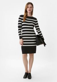 Strickkleid mit schwarz-weißen horizontalen Streifen, Bootsausschnitt, taillierte Silhouette, kombiniert mit einer schwarzen Handtasche und quadratischen Absatzschuhen.