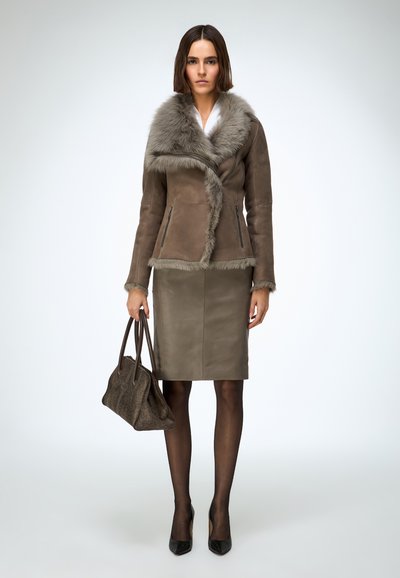 Blouson en shearling marron avec col en fourrure grise, coupe ajustée, associé à une jupe crayon en cuir marron et des escarpins noirs, tenant un sac à main texturé.