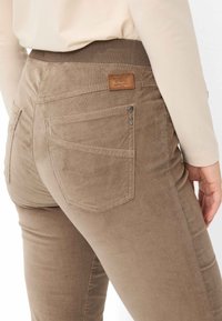 Brown corduroy pants with a stretchy waistband, featuring a leather label, two back pockets, and small metallic accents near the seams.