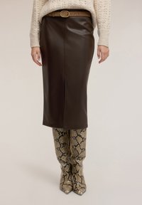 Brązowa spódnica ołówkowa z ekoskóry z przednim szwem, zestawiona z kozakami sięgającymi do kolan o beżowym wzorze skóry węża i zaostrzonych noskach.