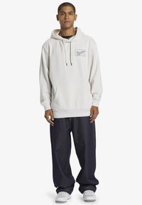 Cream-colored hoodie with a front pocket, drawstring hood, and blue logo patch; paired with loose dark denim jeans and white sneakers.
