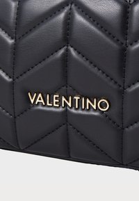 Mustan nahkainen käsilaukku, jossa on tikattu chevron-kuvio, ja jossa on kohokuvioitu "Valentino" -logo kullatussa metallissa. Teksturoidut pinnat ja selkeät linjat.