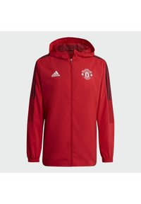 Chaqueta ligera roja con capucha, cierre de cremallera, acentos en negro a rayas en los hombros y logo del Manchester United en el lado izquierdo del pecho.